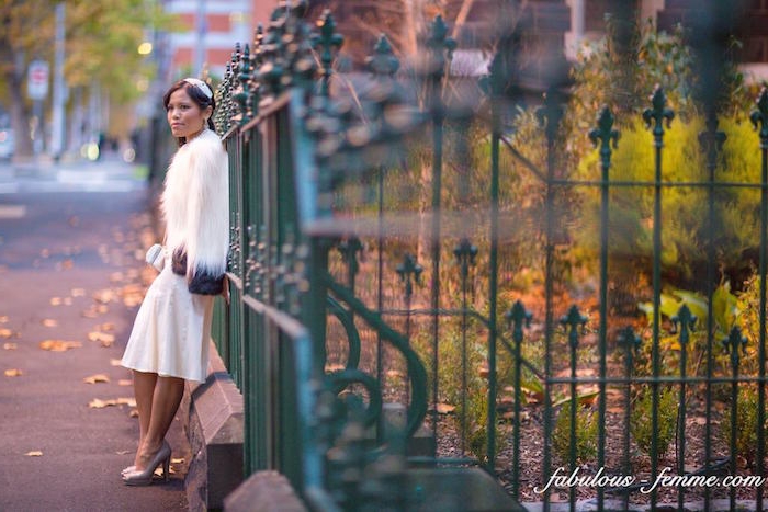 fluffy white fur coat, worn over a white silky midi dress, by a brunette woman, with a small white hat, and nude colored pumps, leaning on a green, decorative metal fence