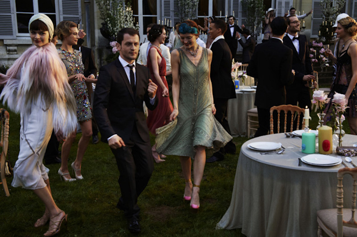 dancing people dressed in roaring 20s fashion, on a green lawn near a house, set table in one corner, woman in a green, embroidered flapper dress, man in a smocking