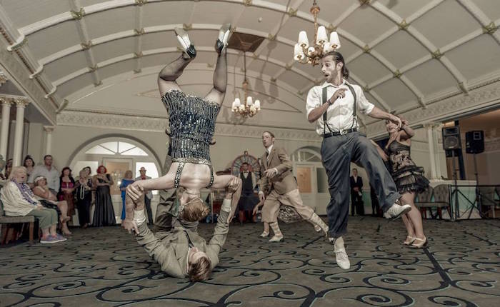 hall with a retro ornamental carpet, and a decorated ceiling, people dressed in roaring 20s fashion, dancing and flipping int he air