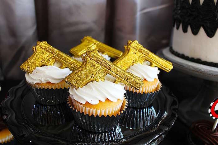 four vanilla cupcakes, with white frosting, topped with small, sparkling gold gun figurines, placed on a black dish, 60th birthday party ideas for men, golden gun themed bond party 