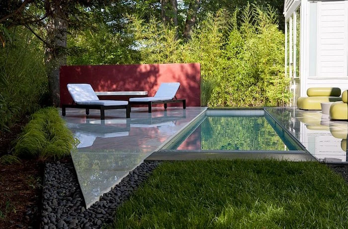 sun beds in white, and a matching table, on a smooth triangular panel, placed in a garden, near a rectangular pool, small backyard pool ideas, grass and pebbles