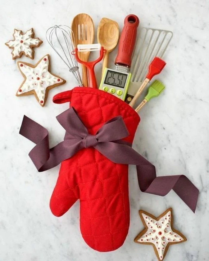 thermometer and wooden spoons, a whisk and a peeler, a spatula and other kitchen utensils, inside a red glove, tied with a purple bow, creative gift ideas, three glazed cookies nearby