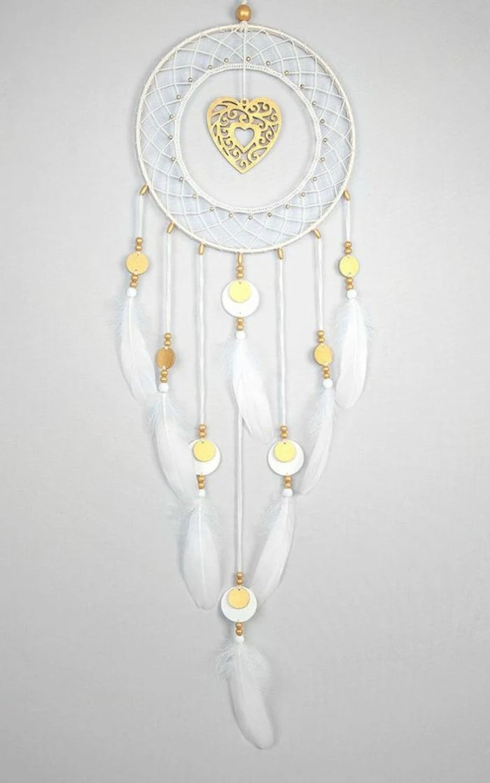 heart ornament in gold, in the middle of a white dream catcher, with white feathers, and more small gold ornaments