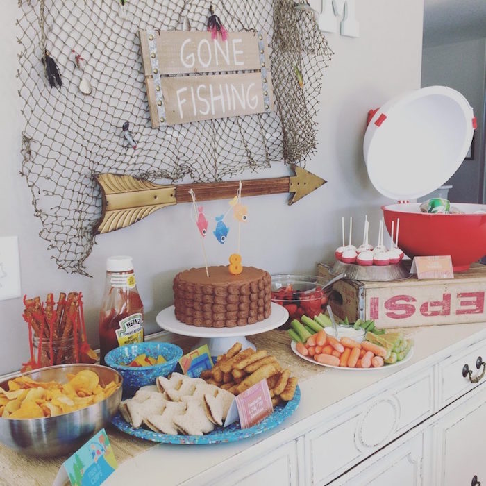 chips sandwiches and other treats, on a table containing a chocolate cake, decorated with fishing themed ornaments, 60th birthday ideas, fishing net and a sign saying gone fishing