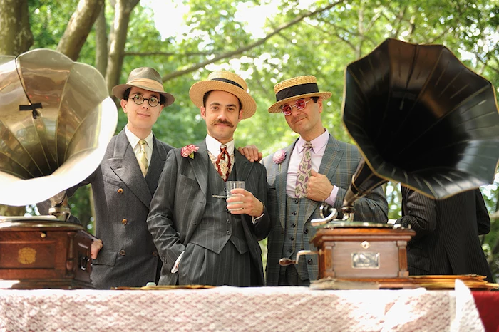 three men in great gatsby outfits, grey three-piece patterned tweed suits, colorful ties and straw hats, two antique gramophones nearby
