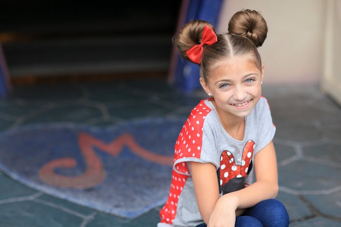 little girl haircuts, symmetrical hair buns, on the head of a smiling, brunette young girl, wearing a minnie mouse t-shirt, and a single red bow