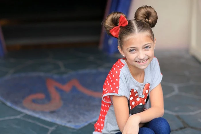 little girl haircuts, symmetrical hair buns, on the head of a smiling, brunette young girl, wearing a minnie mouse t-shirt, and a single red bow