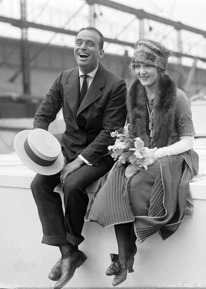 roaring 20s dress, on a smiling woman, with a fur shawl and a hat, sitting next to a laughing man, in a vintage suit, authentic antique photo