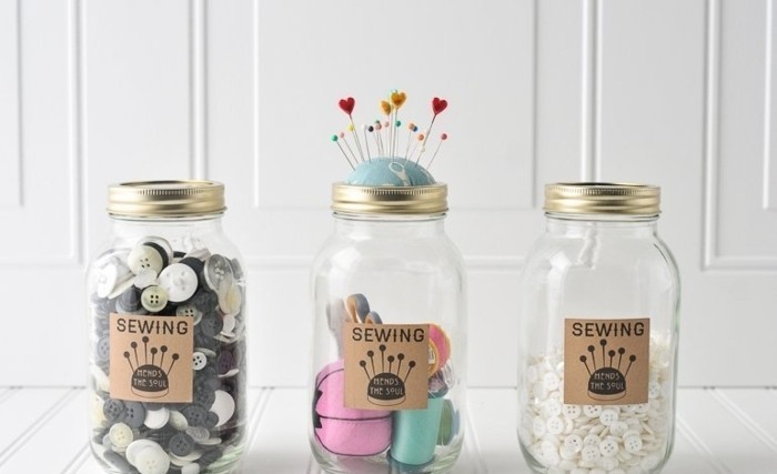 sewing accessories organizers, made from mason jars, with beige labels, two containing buttons, and one thread and pins