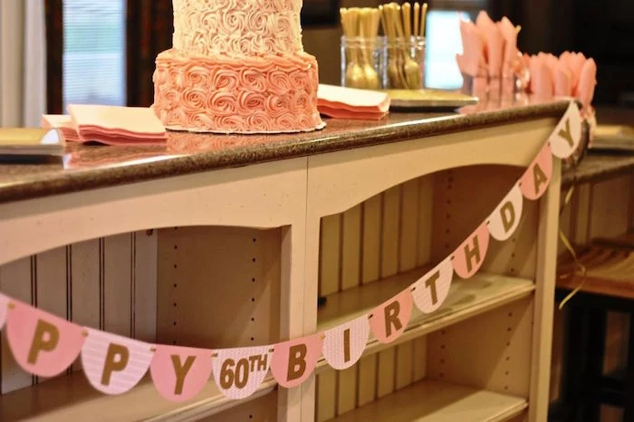 garlands in two shades of pink, decorated with the inscription happy 60th birthday, hung on a wooden counter, with a pink birthday cake, gold cutlery and pink napkins