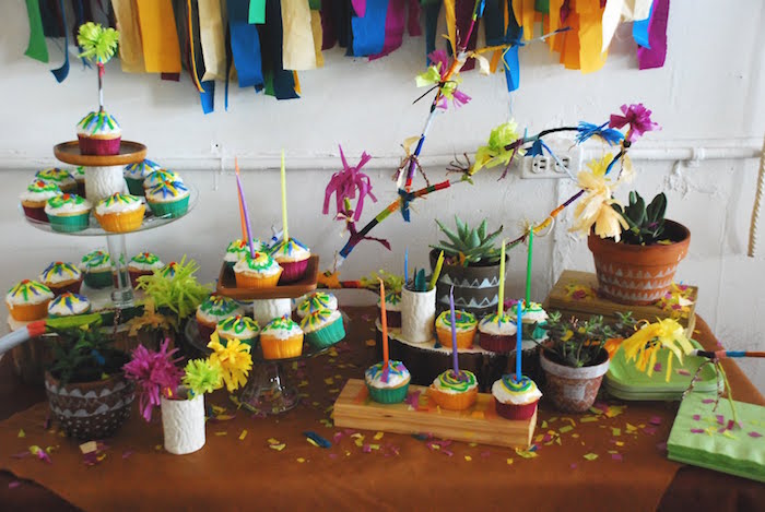 confetti and paper garlands in different colors, decorating a table, covered with multicolored cupcakes 60th birthday color, brown tablecloths and potted succulents