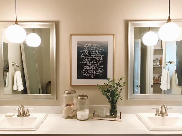 symmetrical bathroom setup, featuring two sinks, two lit lamps, and two mirrors, nice bathrooms, framed poster with writing in white, and two jars with toiletries