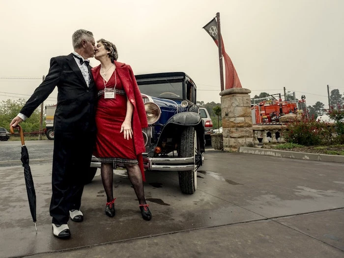 elderly couple dressed in 1920s inspired clothes, kissing in front of a black antique car, red dress and black smocking