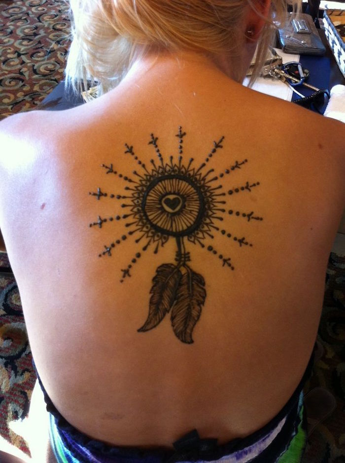 blonde woman with hair tied up, showing her bare back to the camera, it is decorated with a dreamcatcher, painted with dark brown henna