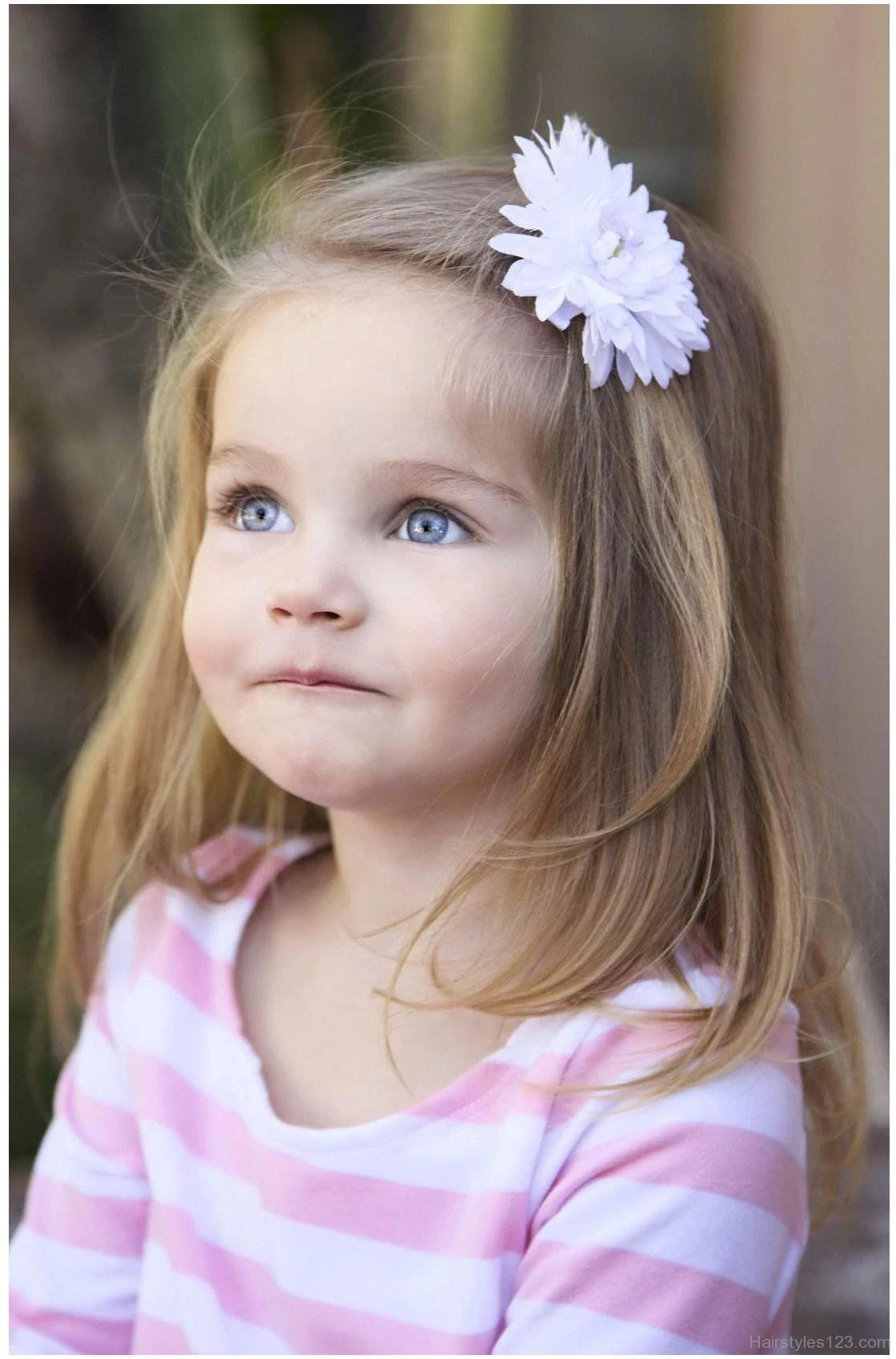 close up of a young child, with ash blonde, shoulder-length hair, decorated with a white flower ornament, cute girls hairstyles, white and pink striped jumper