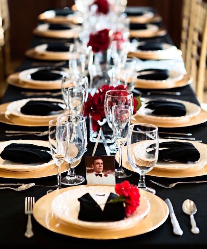 napkins in black and white, folded to look like tuxedos, on a long black table, set for dinner, 60th birthday color, white and black theme, decorated with red flowers, and a photo of sean connery
