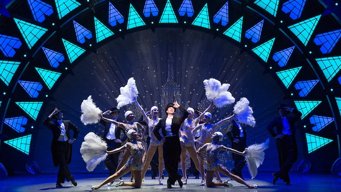 female dancers holding ostrich fans, and male dancers, wearing smockings and top hats, posing on an elaborately decorated stage, bob crowley