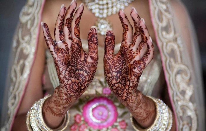 deep red henna patterns, on the palms of the hands of an indian bride, wearing a gown, with pink and gold embroidery