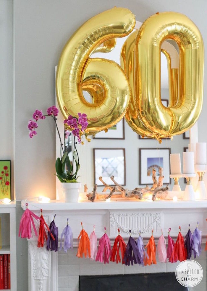 large balloons in gold, shaped like the numbers 6 and 0, near a mantelpiece, decorated with streamers in pink, red and violet