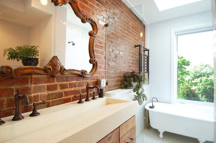 brickwork wall with a large mirror, in an antique ornate frame, a clawfoot bathtub, and the sink with two antique faucets, bathroom wall decor ideas, large window and potted plants
