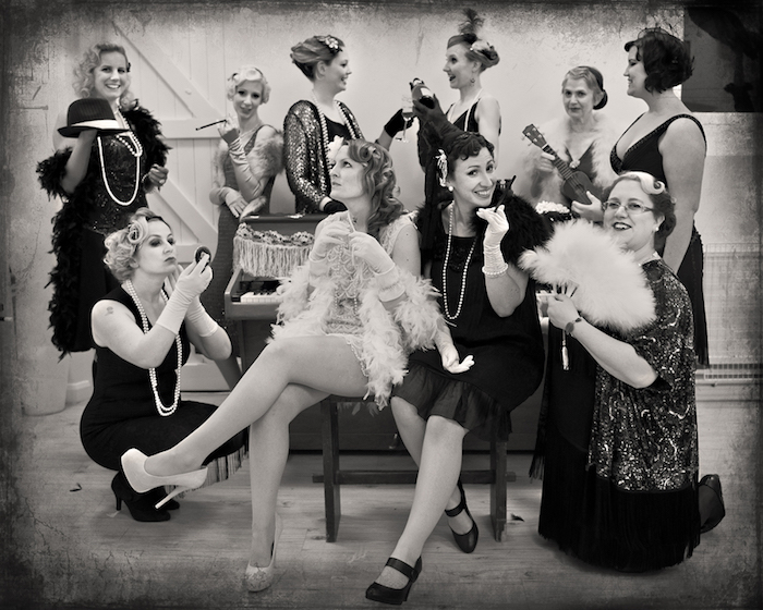 greyscale faux vintage photo, showing a group of women, all dressed in gatsby inspired dresses, with feather boas, long pearl necklaces, smiling and posing for the camera