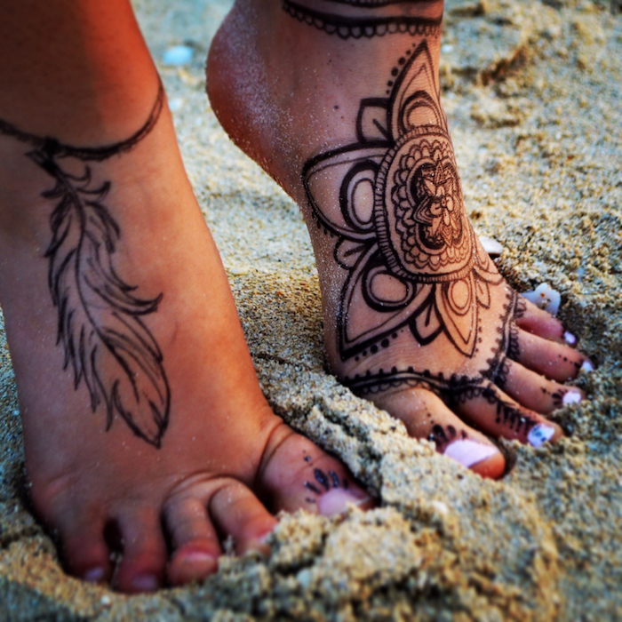 bare feet on a sandy surface, each is decorated with a different henna foot tattoo, one has a feather, and the other a flower