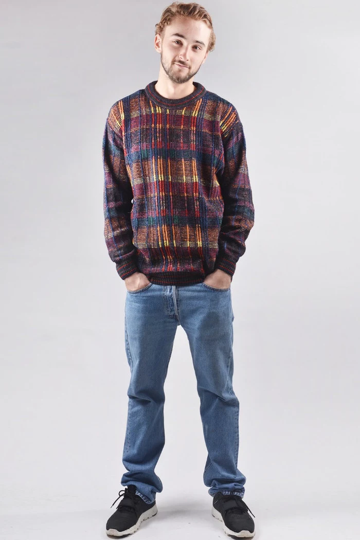 smiling young man, with short mustache and beard, wearing a long, multicolored sweater, black sneakers and baggy jeans