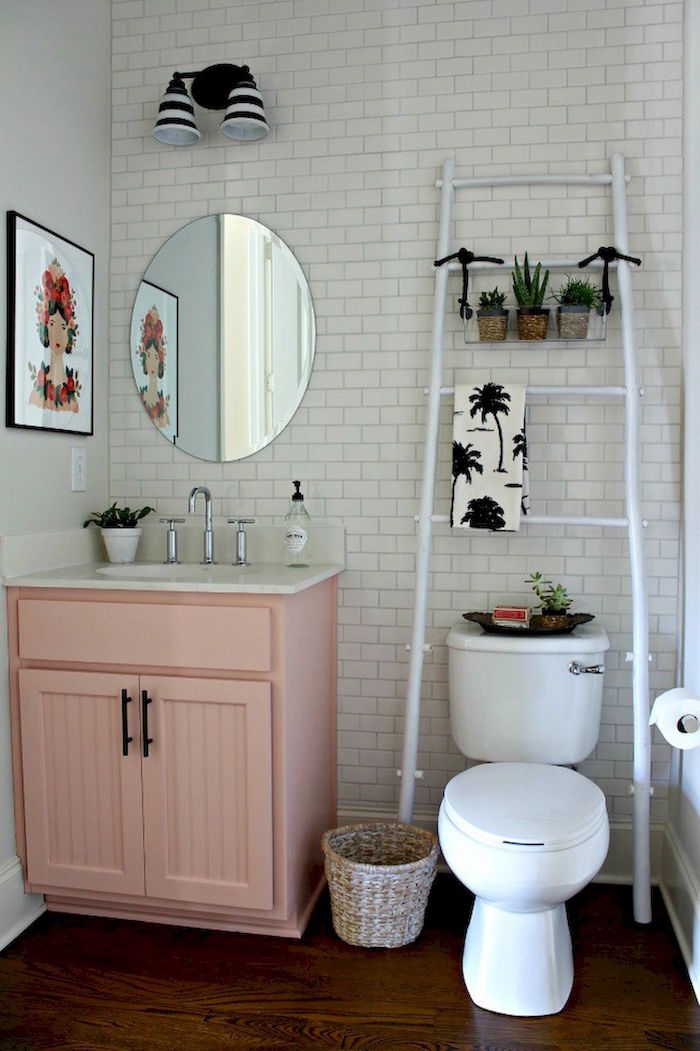 decorative white ladder, with three small potted plants, in a room wit white subway tiles, pale pastel pink cupboard with an inbuilt sink, and a white toilet, small bathroom décor, oval mirror and framed artwork