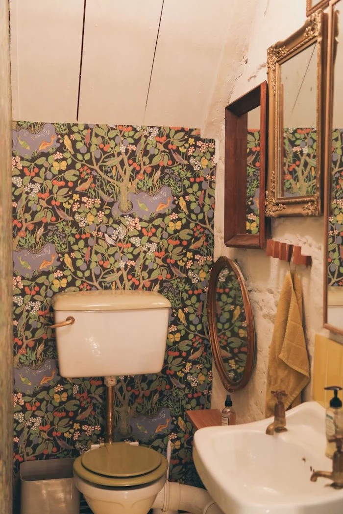 wallpaper in green, depicting trees and birds flowers and fruit, in a small room, with a vintage toilet bowl, a white stink, and several mirrors in wooden frames, small bathroom decoration ideas, floor to ceiling cupboard with white wooden boards