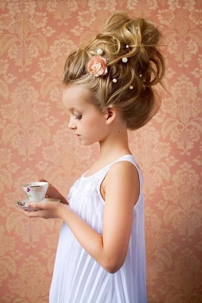 voluminous blonde up-do, decorated with pearl-like details, and a peach floral motif, worn by a small child, holding a teacup, in a formal, white sleeveless dress