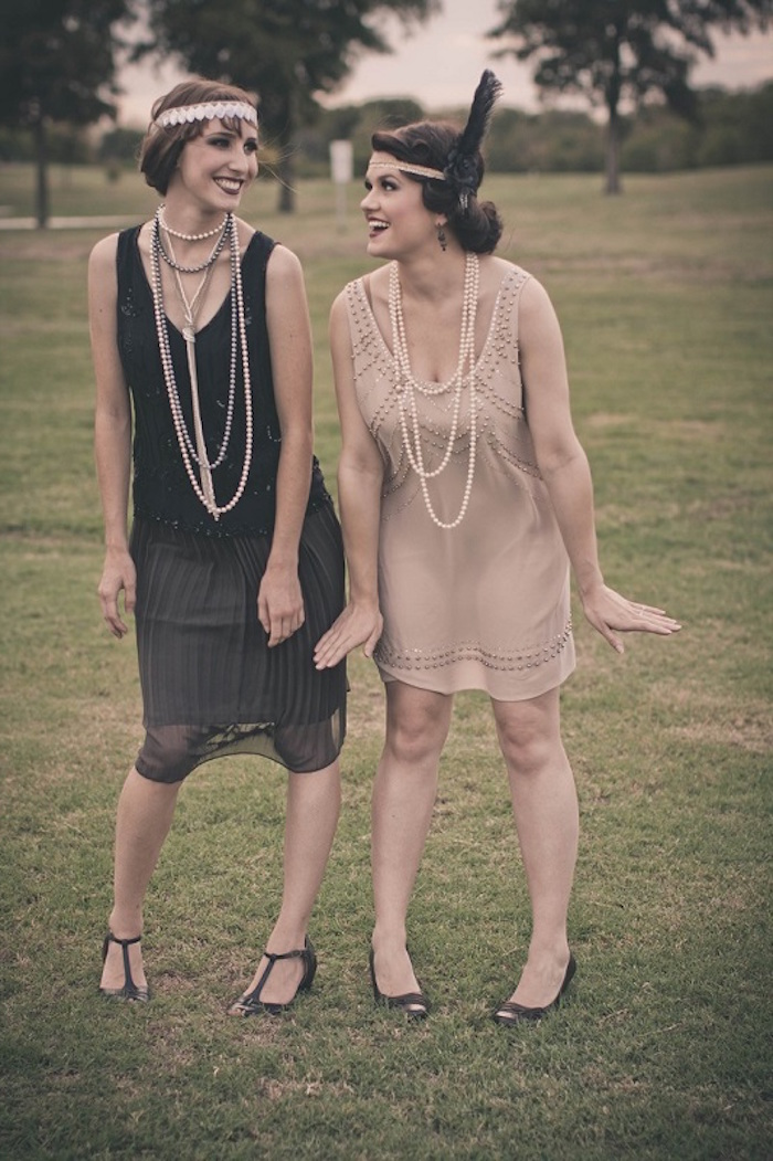 smiling women posing for a photo, one dressed in a midi, black roaring 20s dress, with t-bar shoes, a headband and long pearl necklaces, the other is in a similar dress in pale pink, how to dress up for a roaring 20s party