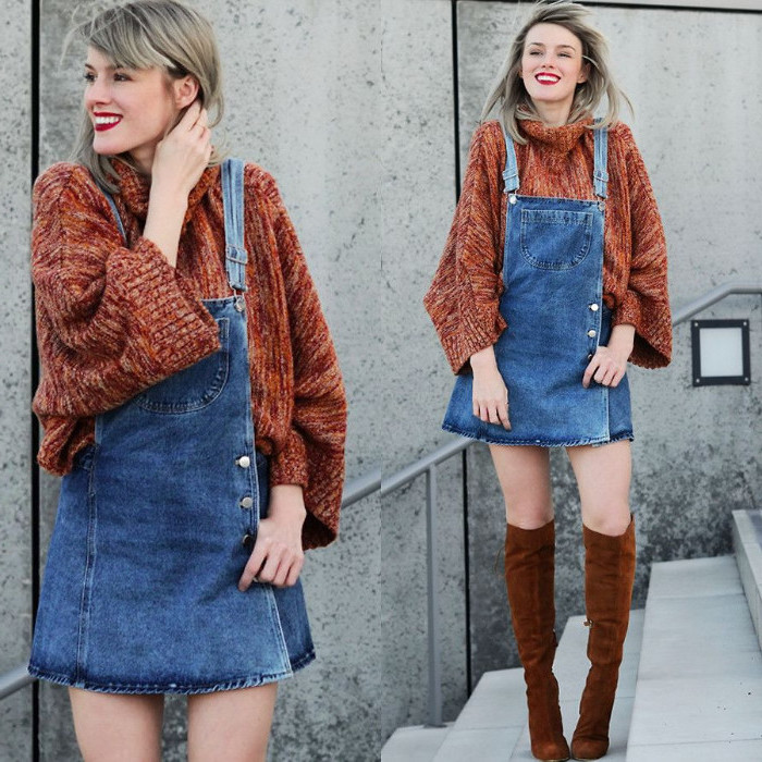 dungaree dress made from denim, with side buttoning, worn over a bulky, brick red turtleneck jumper, by a smiling blonde woman, with knee-high brown suede boots