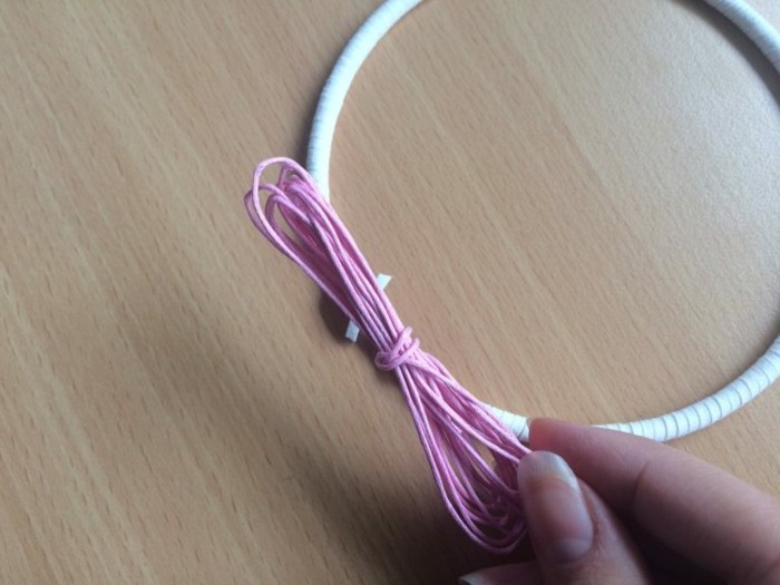 string in pink, held by a hand, near a wire hoop, wrapped in a white leather rope, how to weave a dreamcatcher, wooden surface in the background