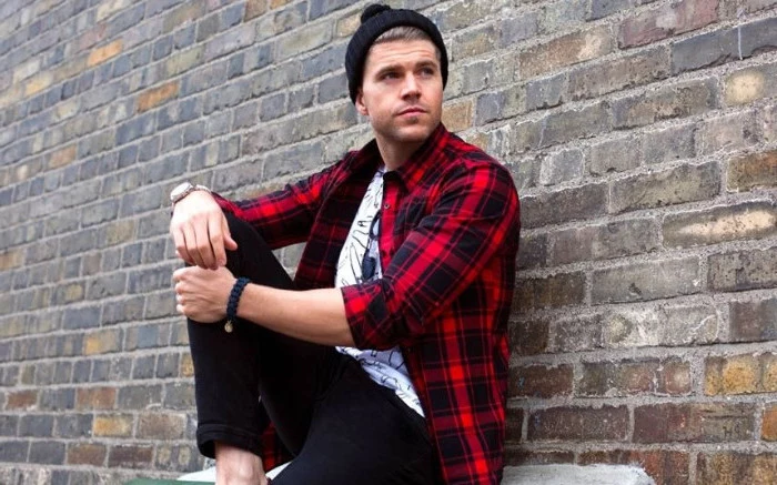 sitting man with black beanie hat, wearing an unbuttoned, red and black plaid shirt, and a white tee with black print, skinny black trousers