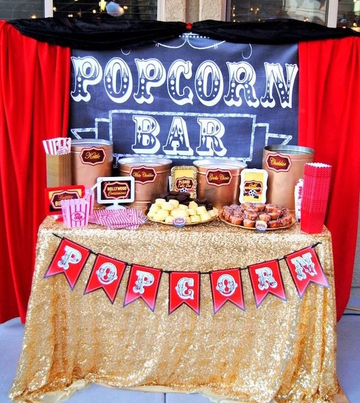 curtains in red and black, around a table with a sign, reading popcorn bar, 60th birthday decorations, a selection of various snack, gold lame tablecloth, and red flags garland 