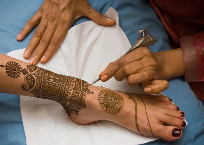 artist drawing henna patterns, on the ankle and foot of a woman, wearing dark cherry red nail polish