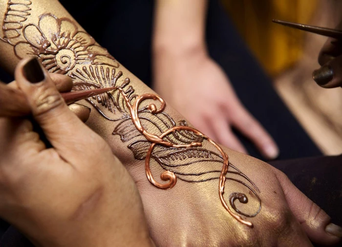 hand with black nail polish, applying gold-colored paint, on top of the floral motifs, of a henna tattoo 