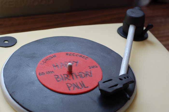 party cake shaped like a vintage record player, with a black and red vinyl record, all made from colorful fondant, happy 60th birthday paul, written In black on top