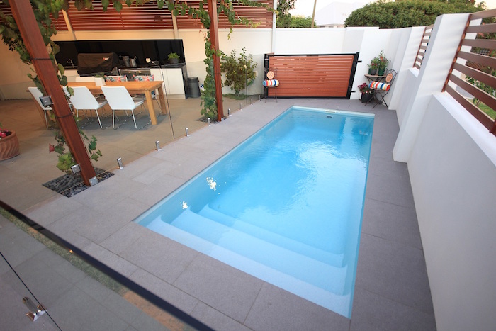 table with six chairs, near a blue rectangular pool, surrounded by grey tiles, small inground pools, with a glass fence 