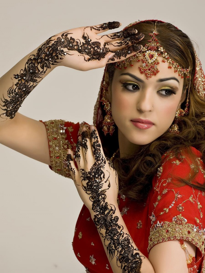 henna tattoo, indian bride in a red gown, embroidered with gold details, and wearing elaborate jewelry, lifting her arms up, to show dark brown mehndi, on her palms and forearms