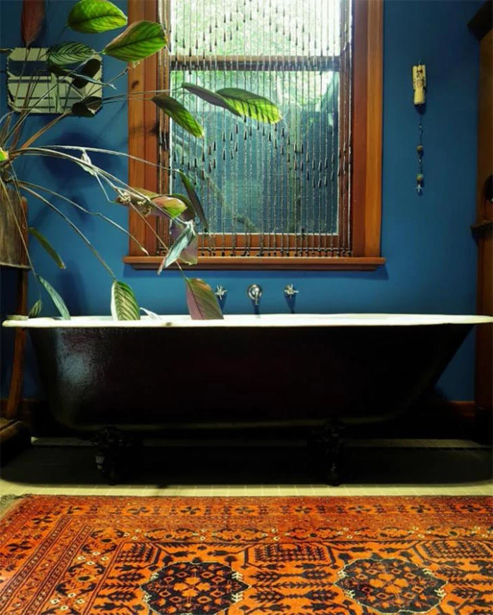 black and white tub, in a room with blue walls, containing a window with a brown frame, a potted plant, and a kilim rug in red, with a dark oriental pattern, diy bathroom