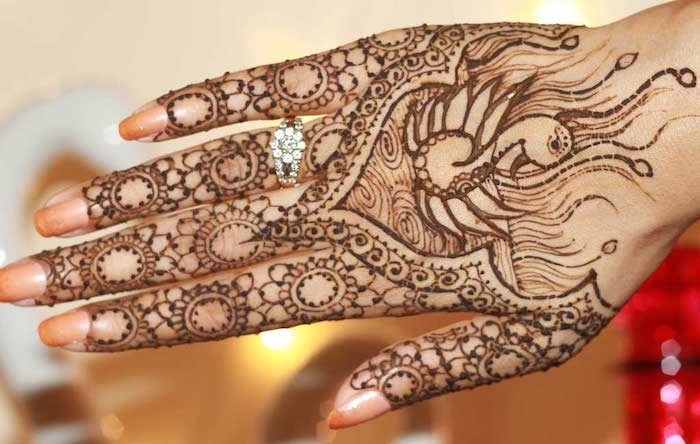 firebird-motif and floral elements, drawn with brown henna, on a hand with pale peach nail polish, and an encrusted ring