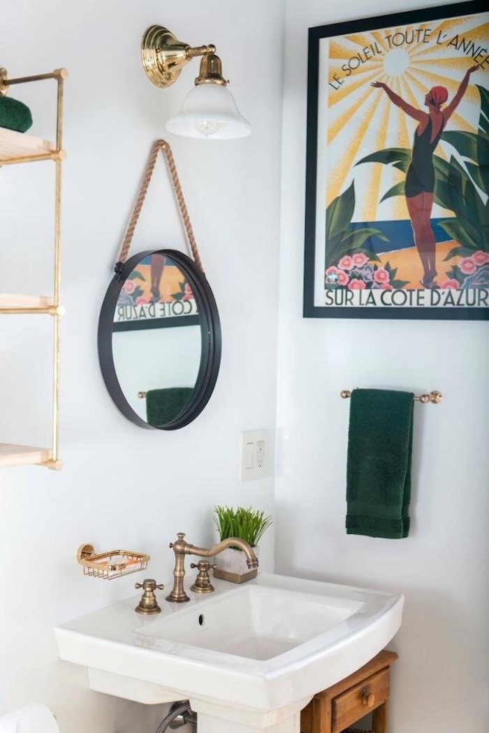 vintage brass faucet, on a white sink, in a room with white walls, decorated with a framed poster, a round mirror, a gold lamp, and other items, small bathroom décor, gold metal shelves