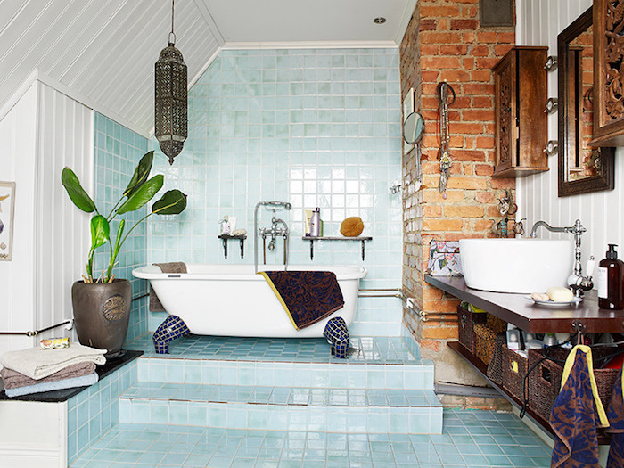 light blue tiles, and white panelling, inside a bright bathroom, containing a tub, a wooden counter with a sink, brickwork detail and engraved wooden cupboards