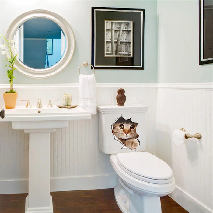funny cat decal sticker, on a white toilet bowl, bathroom decorating ideas on a budget, room with pale duck's egg blue walls, and white panelling, containing a round mirror, and framed black and white images