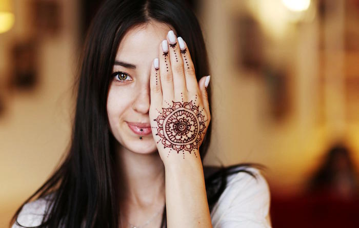 brunette girl with long dark hair, parted in the middle, smiling white covering one of her eyes with her hand, decorated with a mehndi, henna meaning