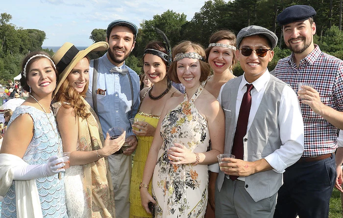 friends dressed in roaring 20s inspired outfits, smiling at the camera, and holding drinks, flapper dressed with hats and headbands, smart trousers with vests and suspenders