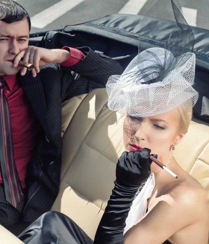 tulle hat in white and black, worn by a woman in black and white dress, smoking a cigarette, great gatsby costumes, in an antique car, next to a man smoking a cigar, what to wear to a gatsby themed party