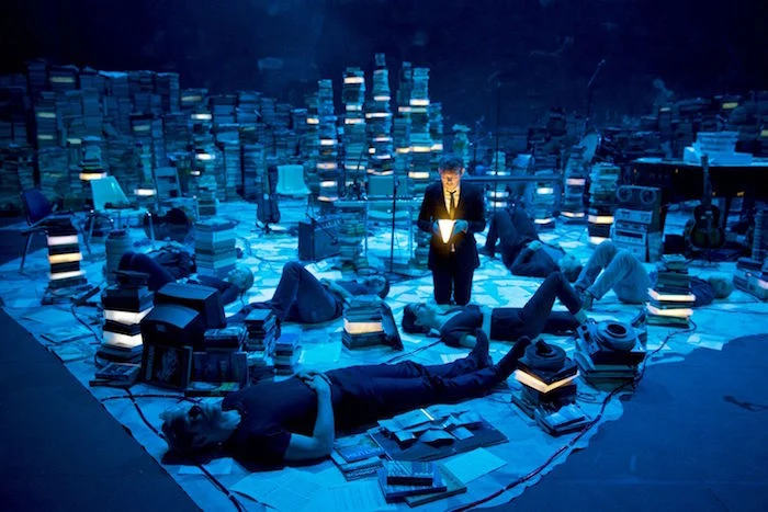 kneeling man in a business suit, holding a glowing book, more men lying on the grown around him, amid stacks of books, bathed in blue light