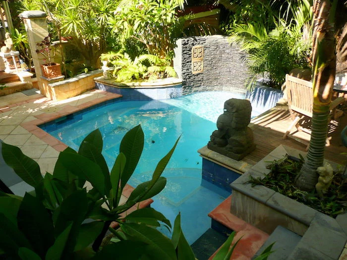 cool backyards, angular pool surrounded by greenery, with a lion statue made of stone, beige and orange tiles on the ground